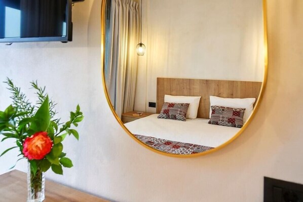 Een slaapkamer met een bed, een spiegel, een vaas met een bloem en een aan de muur bevestigde televisie.