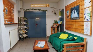 Une pièce chaleureuse avec un canapé vert, une table basse en bois et une bibliothèque. On y trouve une porte sur laquelle est apposée une pancarte, ainsi qu’un panneau d’affichage où sont accrochés divers avis.