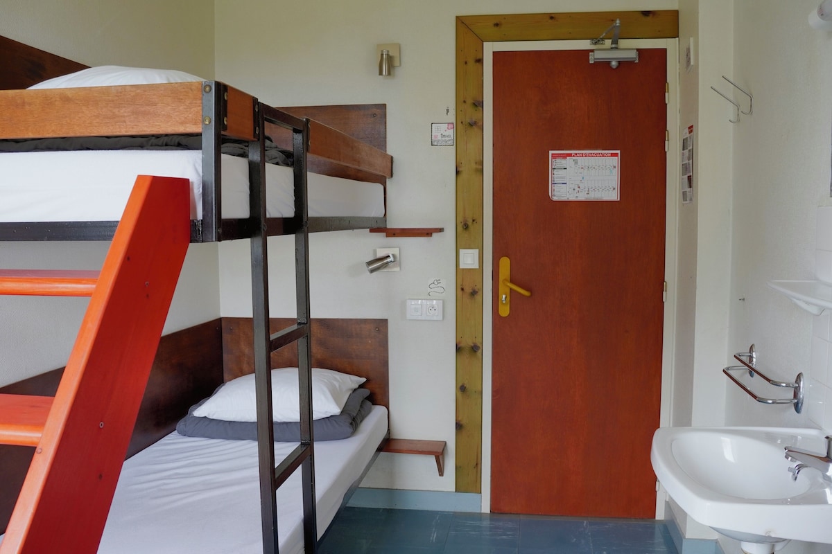 Una cama litera con escalera de madera, una puerta de madera con un letrero, un lavamanos blanco y un piso azul.