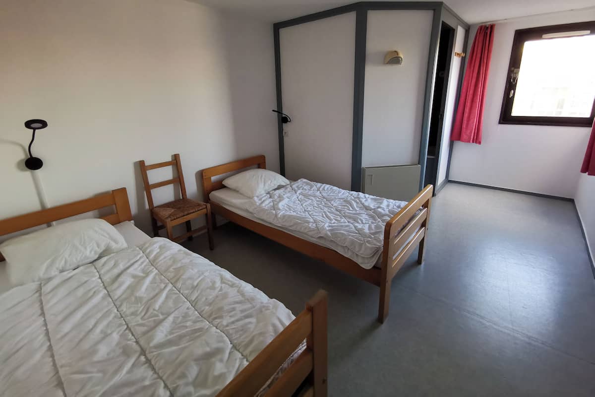 Et værelse med to senge af træ, en stol, et natbord, et vindue med røde gardiner og en fastgjort lampe på væggen.