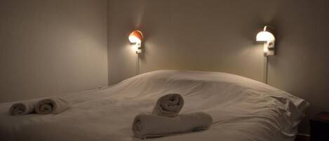 A hotel room with a neatly made bed, two wall-mounted lamps, and rolled towels on the bed.
