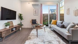 Una sala de estar moderna con un televisor de pantalla plana, un mueble de madera para entretenimiento, un sofá gris, una silla de mimbre, una mesa de centro y un ventanal con cortinas.