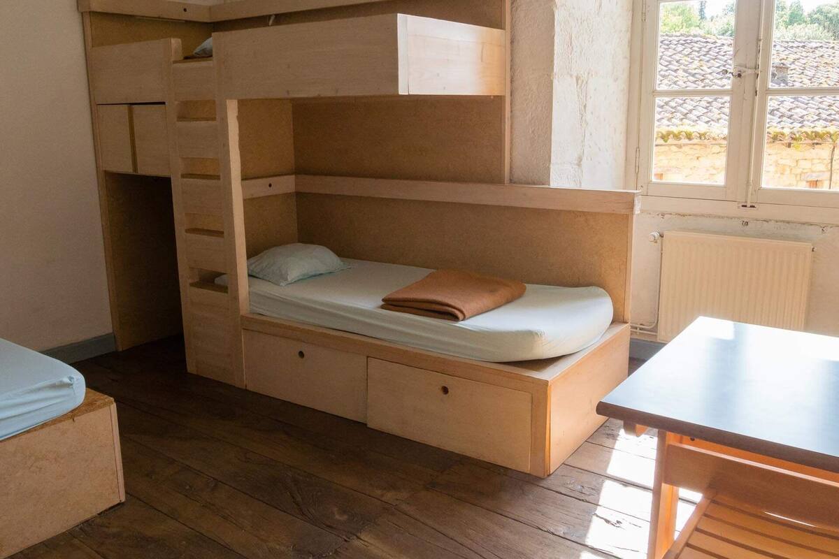 Un lit superposé avec un bureau et des tiroirs, un plancher en bois et une fenêtre donnant sur un toit recouvert de tuiles.