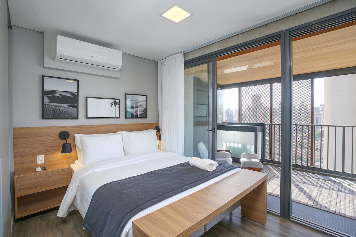 Una habitación de hotel moderna con una cama grande, una mesita de noche, un banco, un balcón con vistas a la ciudad y un aire acondicionado de pared.