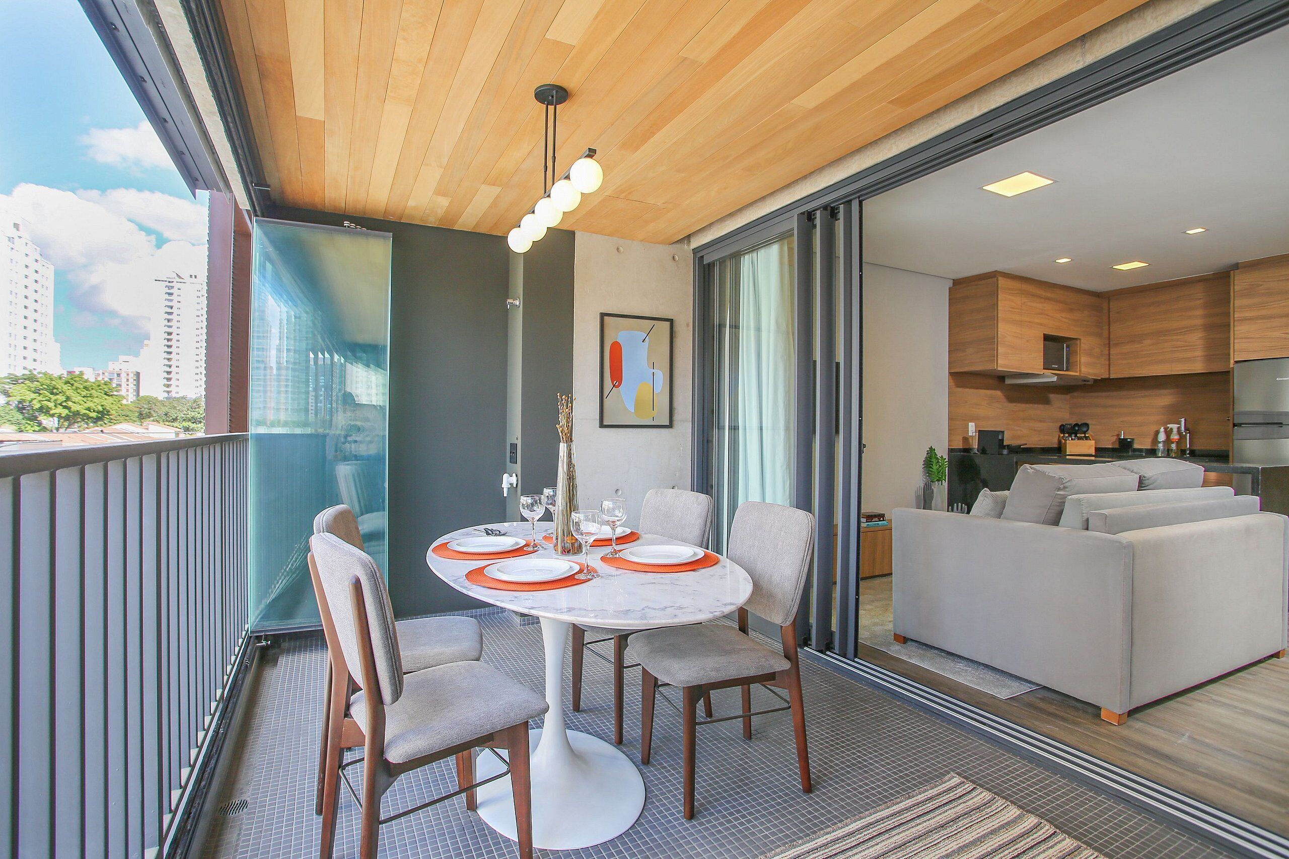 A balcony with a dining table set for four, a sofa, and a kitchen area visible through a glass door.