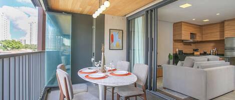 A balcony with a dining table set for four, a sofa, and a kitchen area visible through a glass door.