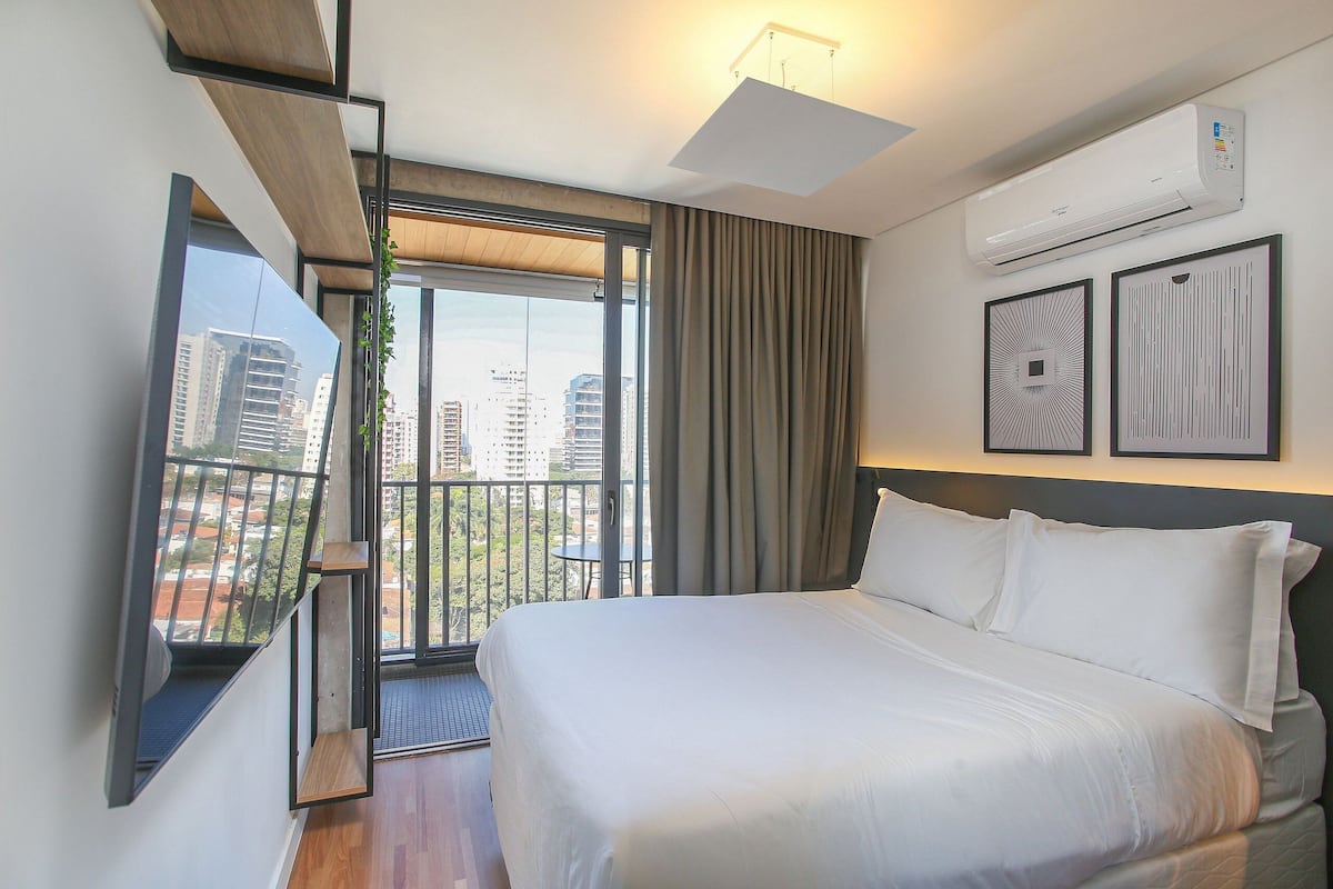 Un hotel moderno con una cama grande, un equipo de aire acondicionado, un balcón con vistas a la ciudad y dos cuadros enmarcados en la pared.
