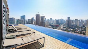 Una piscina infinita en la azotea con sillones de descanso, un horizonte urbano al fondo y un cielo despejado.