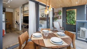 Un comedor con una mesa redonda de madera dispuesta para cuatro personas, un televisor al fondo y una decoración grande de color verde en la pared.