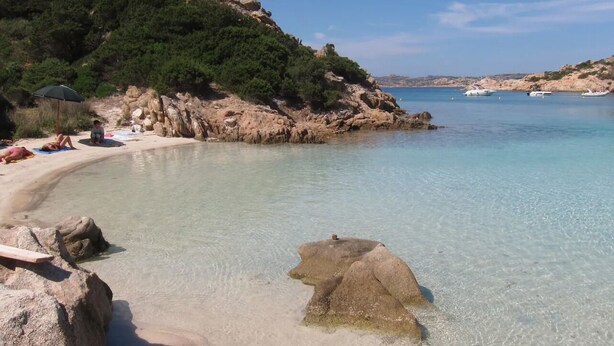 Una playa con agua cristalina, costa rocosa y barcos a lo lejos.