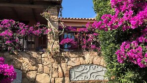 Un muro de piedra con una reja metálica decorativa, un sendero de piedra que lleva a un edificio con techo de tejas, y coloridas flores de buganvilla rosa.