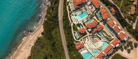 An aerial view of a coastal resort with a large swimming pool, multiple guest villas, and a beach area.
