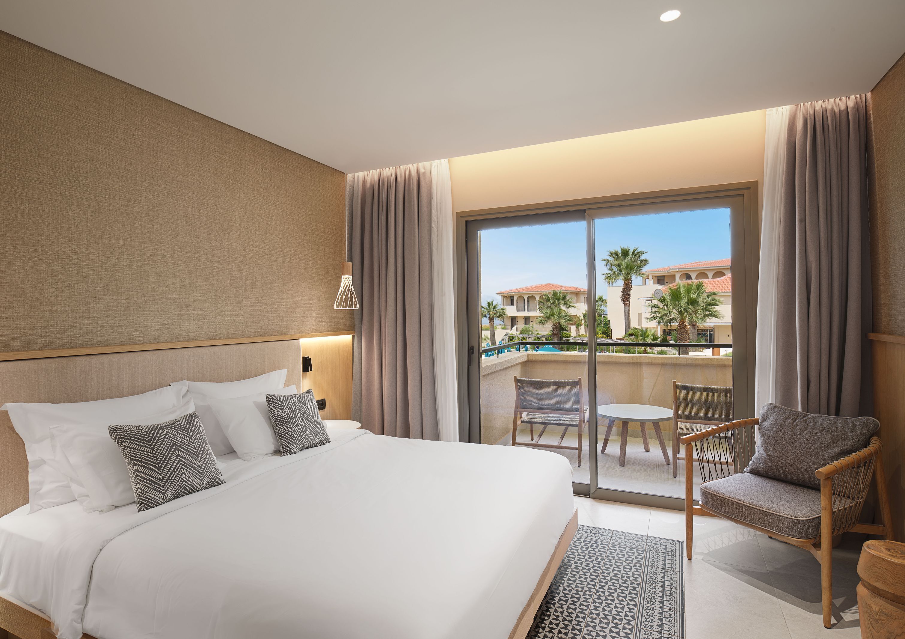 Une chambre d’hôtel avec un lit, une chaise, une petite table, un balcon donnant sur des palmiers et une piscine.