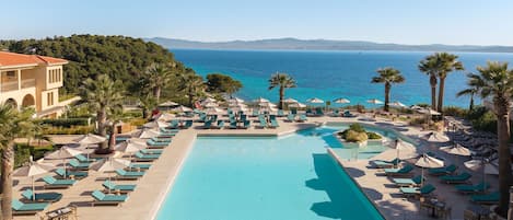 A resort pool with lounge chairs and umbrellas, surrounded by palm trees and a view of the sea.