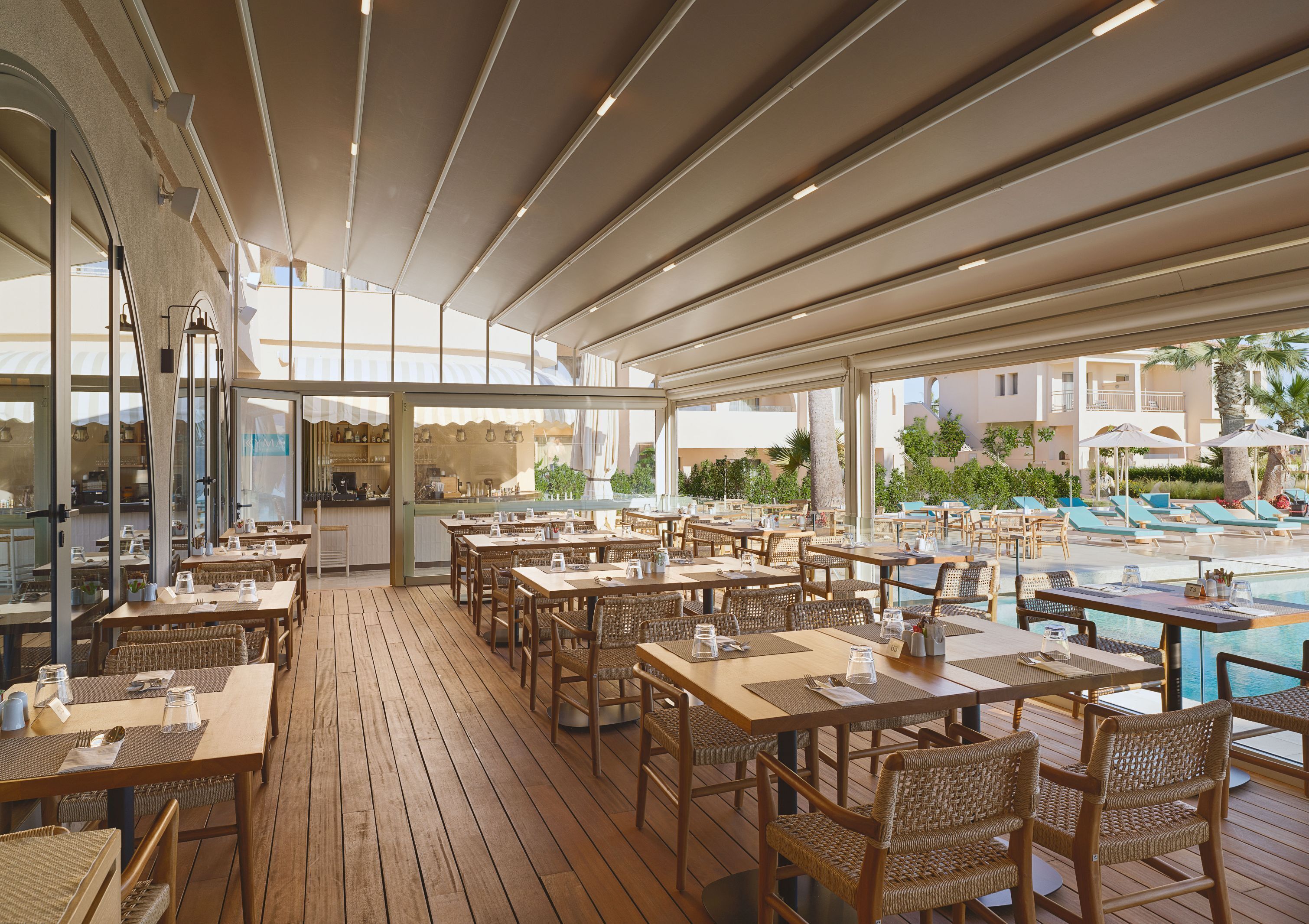 An outdoor dining area with wooden tables and chairs, a poolside lounge, and a view of the pool and buildings in the background.