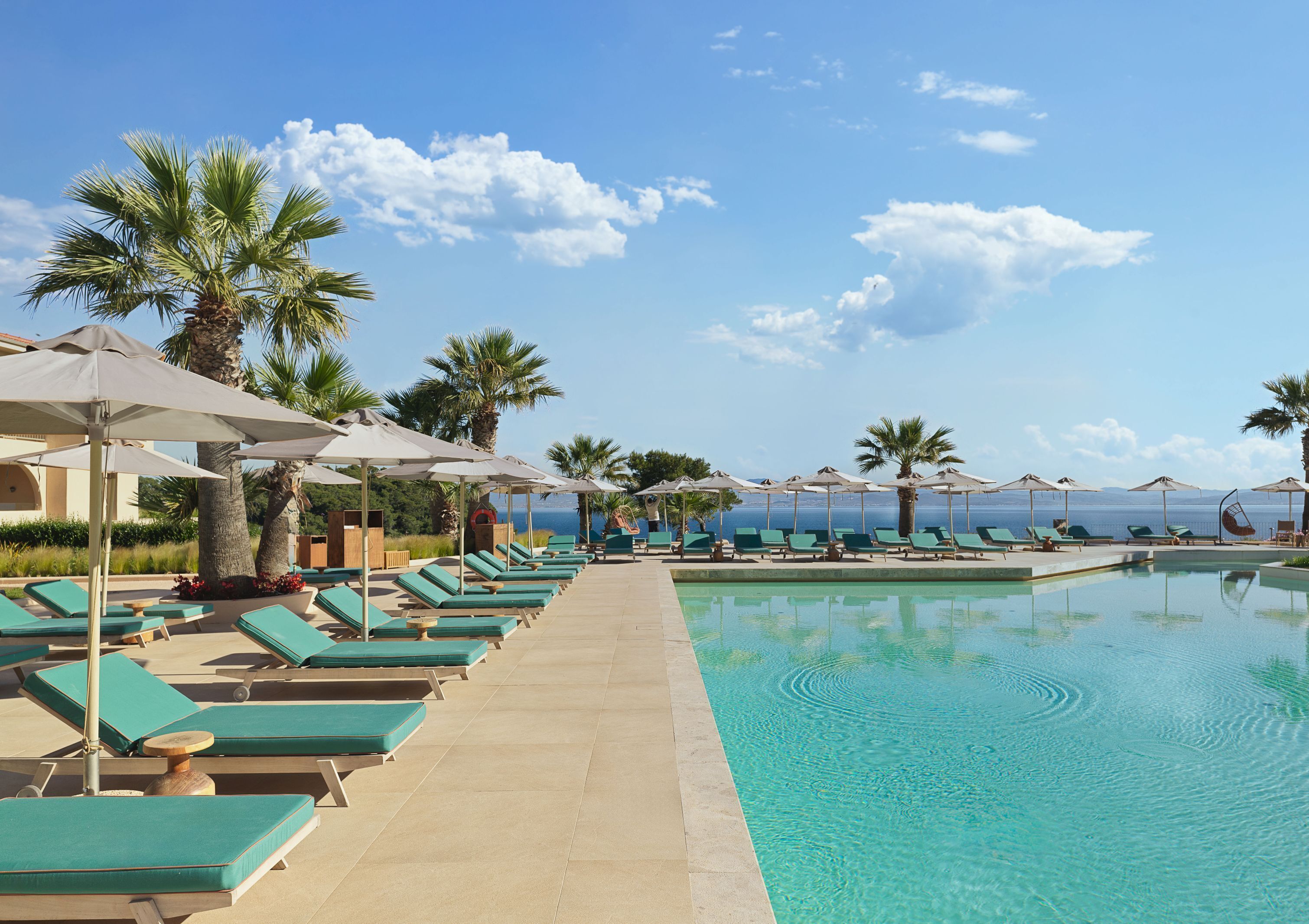A poolside area with lounge chairs and umbrellas, palm trees, and a clear view of the sea.