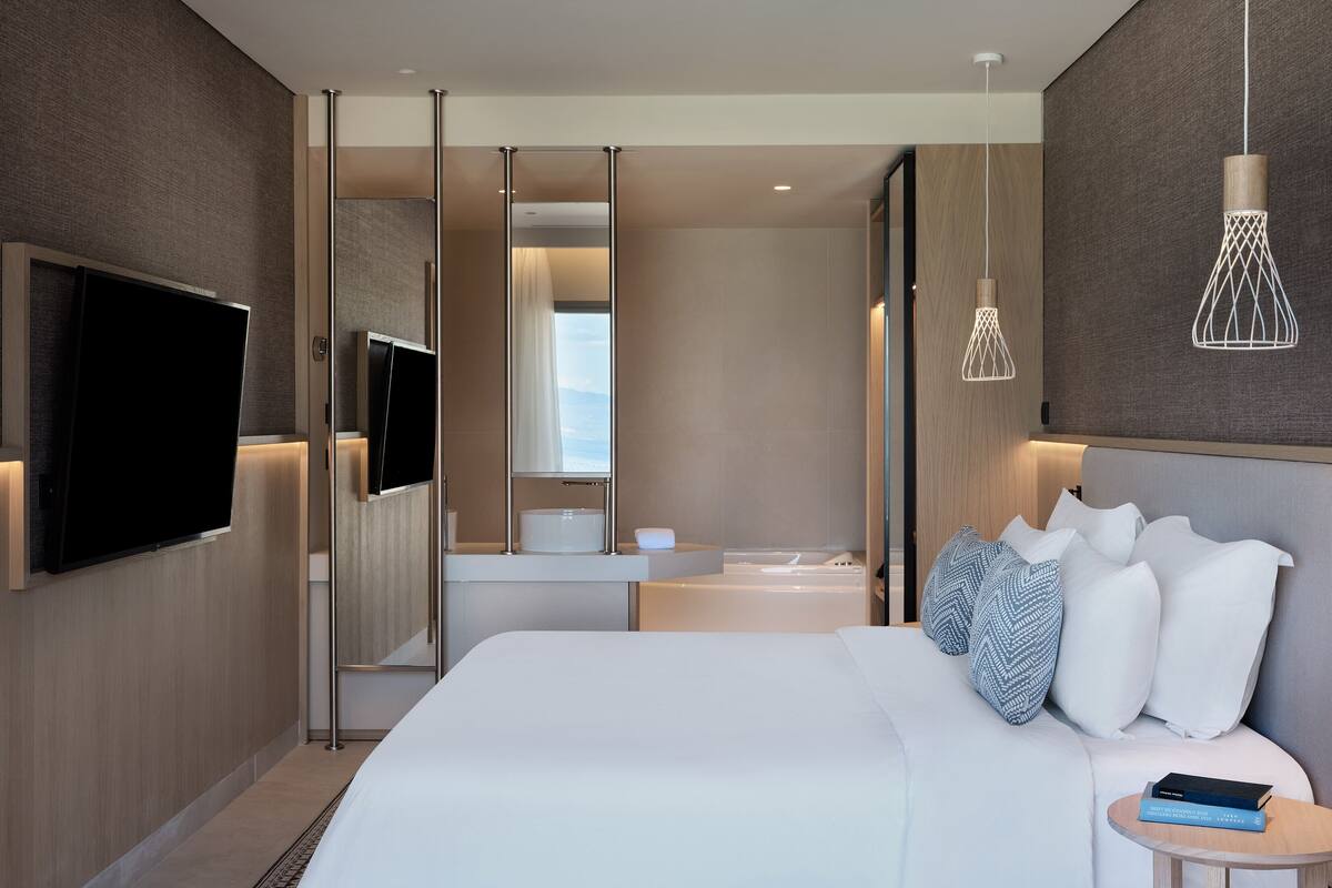 A modern hotel room with a large bed, a TV mounted on the wall, a bedside table with a book, and a view through a sliding door.