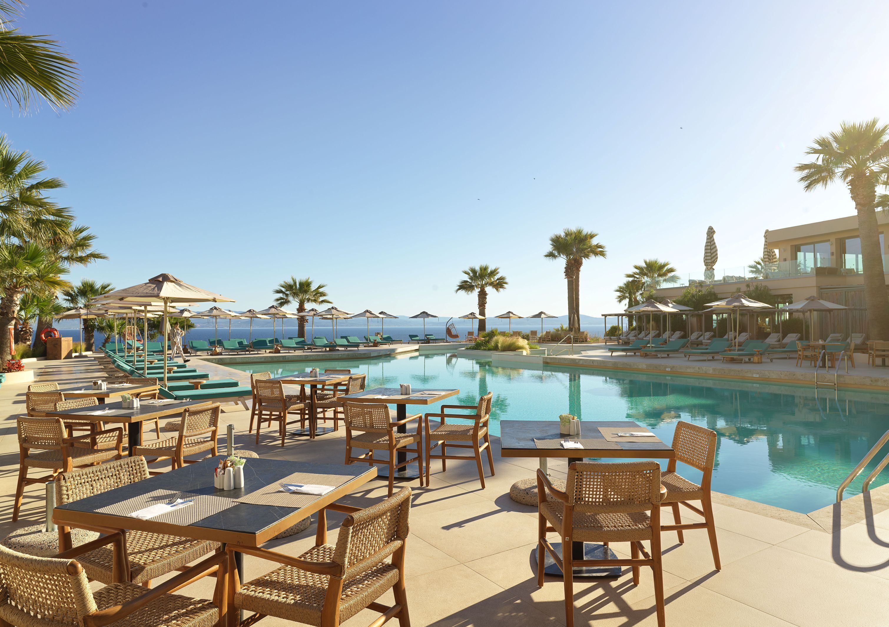 Zona de piscina con sillones, mesas y sombrillas, con vista a un cielo azul despejado y un mar tranquilo.