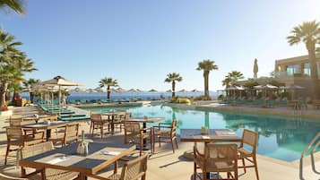 Zona de piscina con sillones, mesas y sombrillas, con vista a un cielo azul despejado y un mar tranquilo.