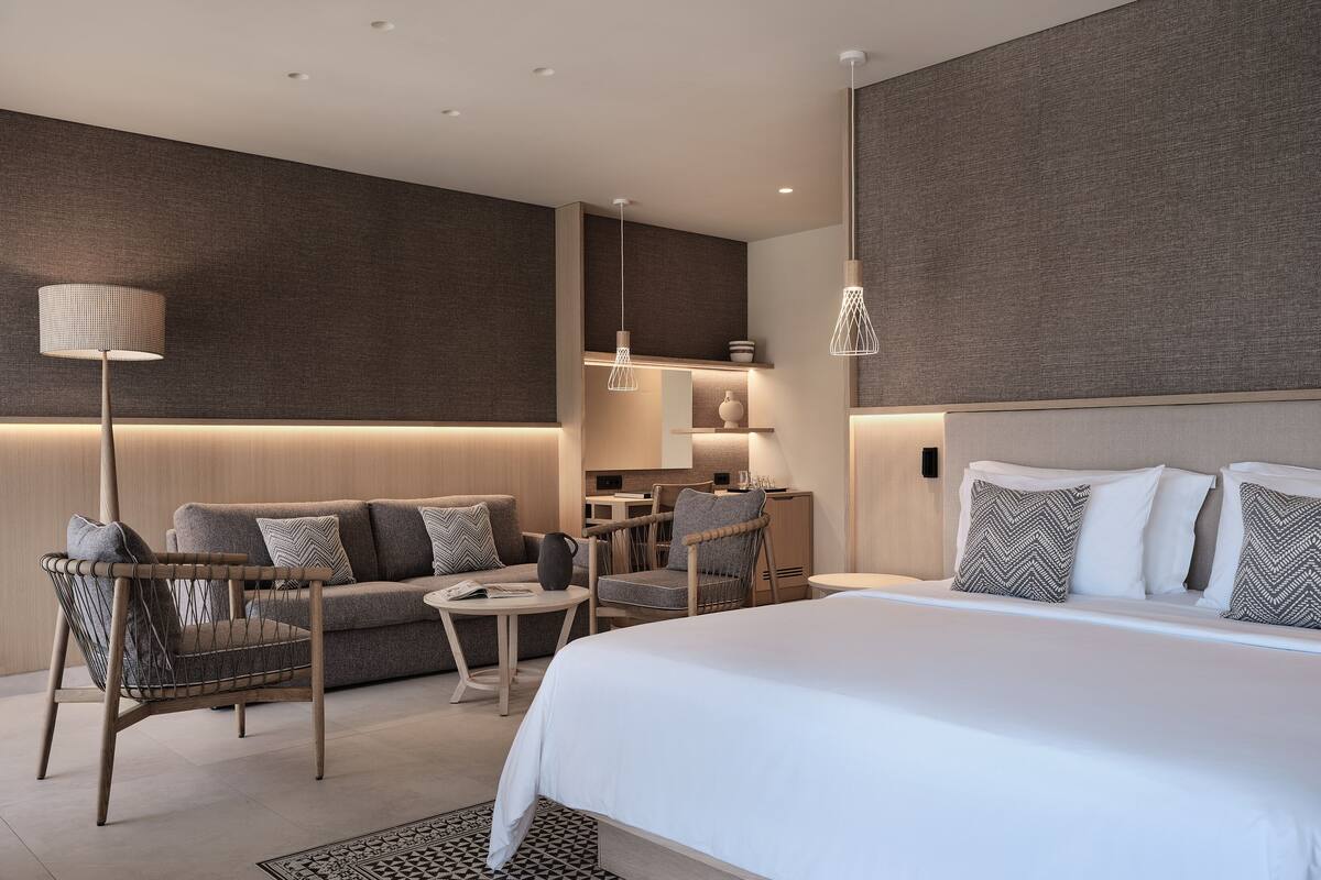 A modern hotel room with a large bed, a sofa, a dining area, and a kitchen.