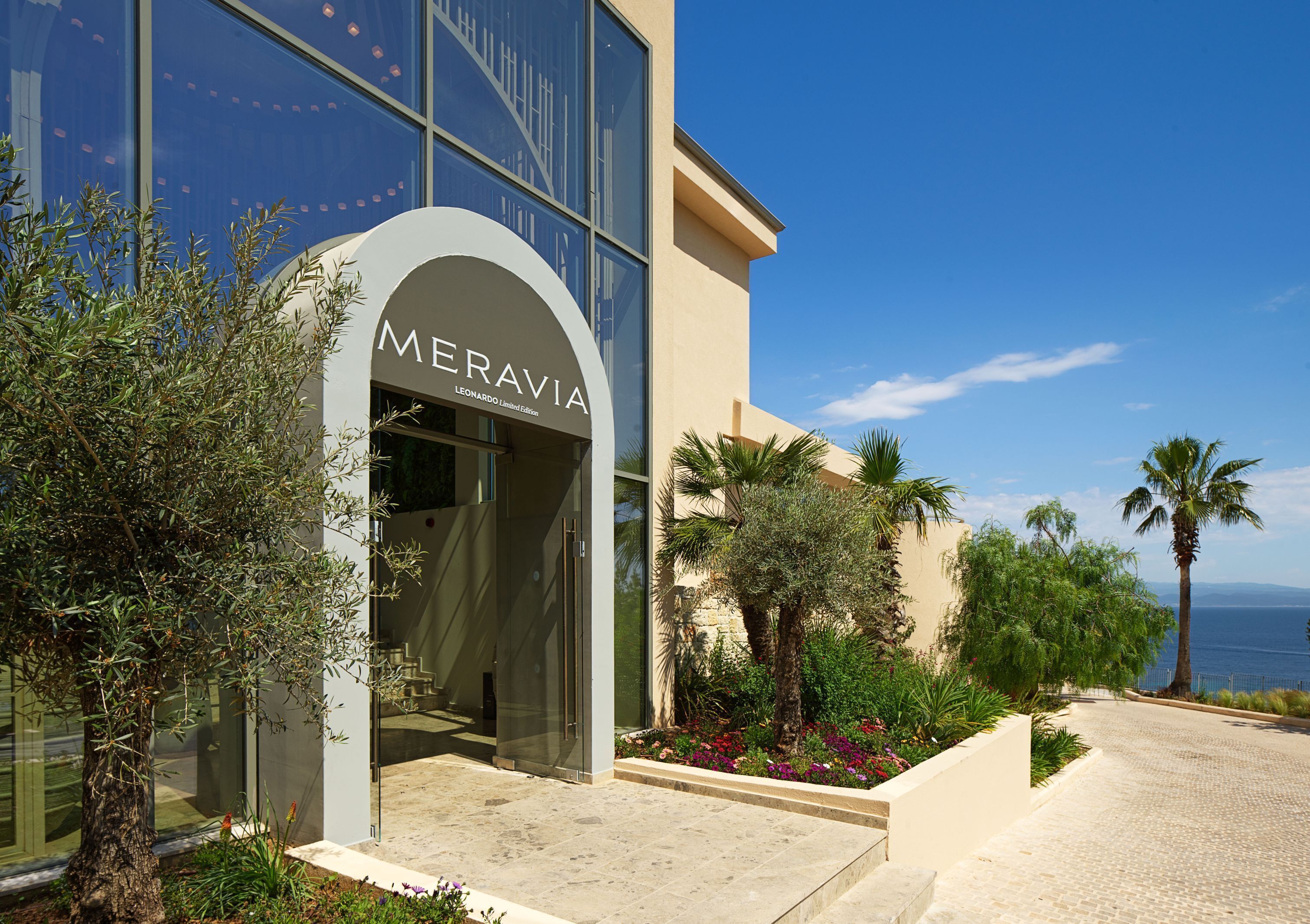 A building with a sign reading 'Meravia Leonardo Limited Edition,' surrounded by greenery and a view of the sea.