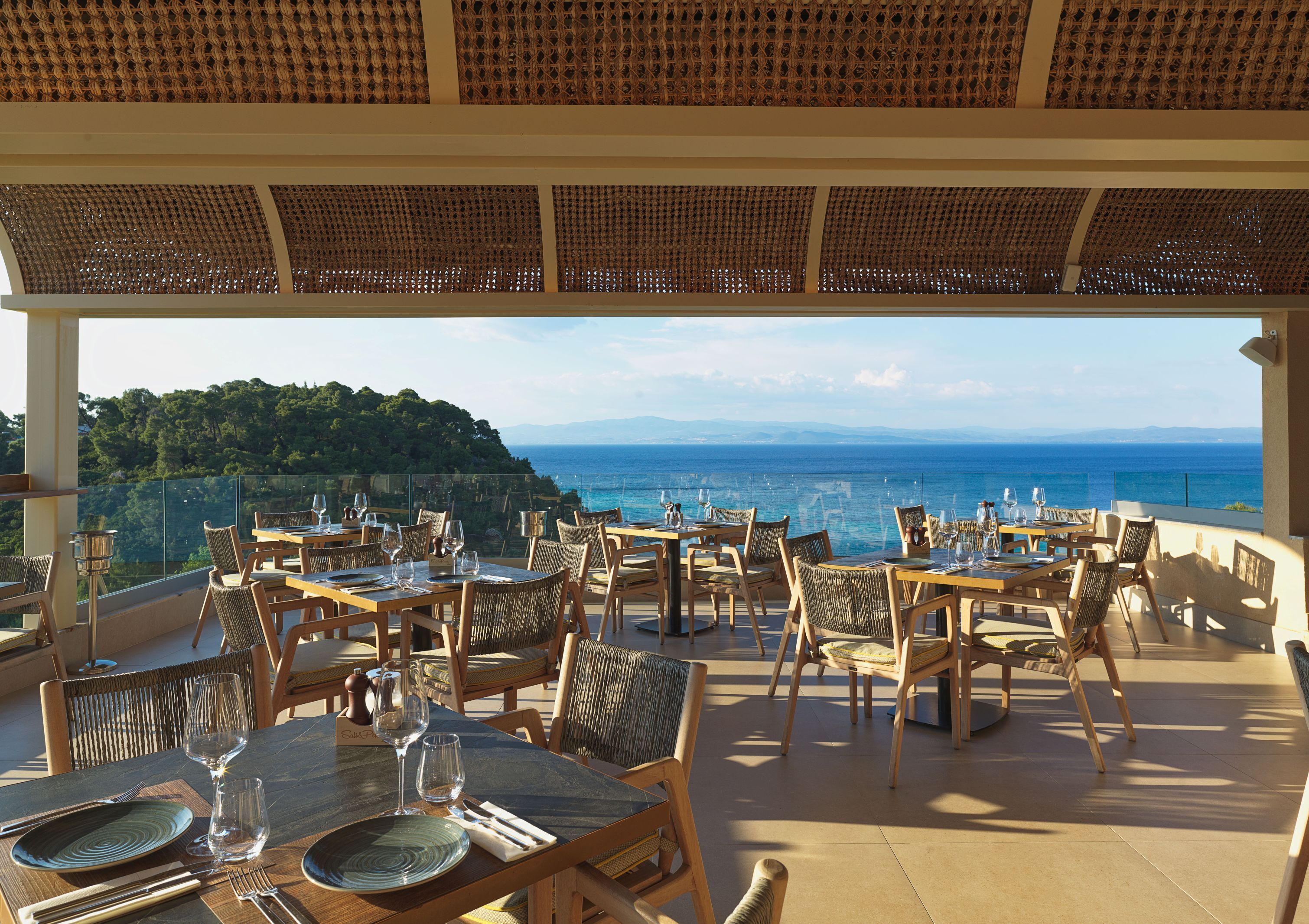 An outdoor dining area with tables and chairs, overlooking a scenic view of the sea and hills.