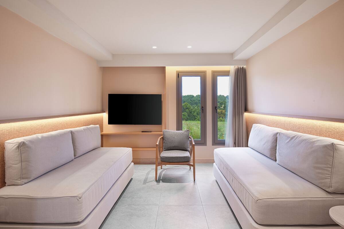 A modern hotel room with two sofas, a TV, and a window with a view of greenery.