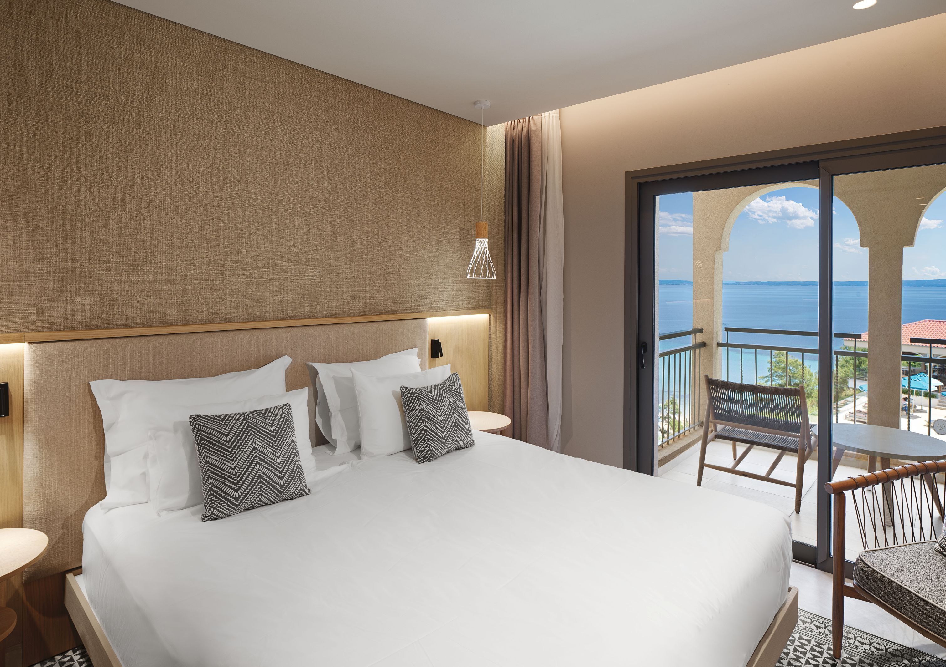 A hotel room with a large bed, two bedside tables, a nightstand, a balcony with a view of the sea, and a small table with a chair.