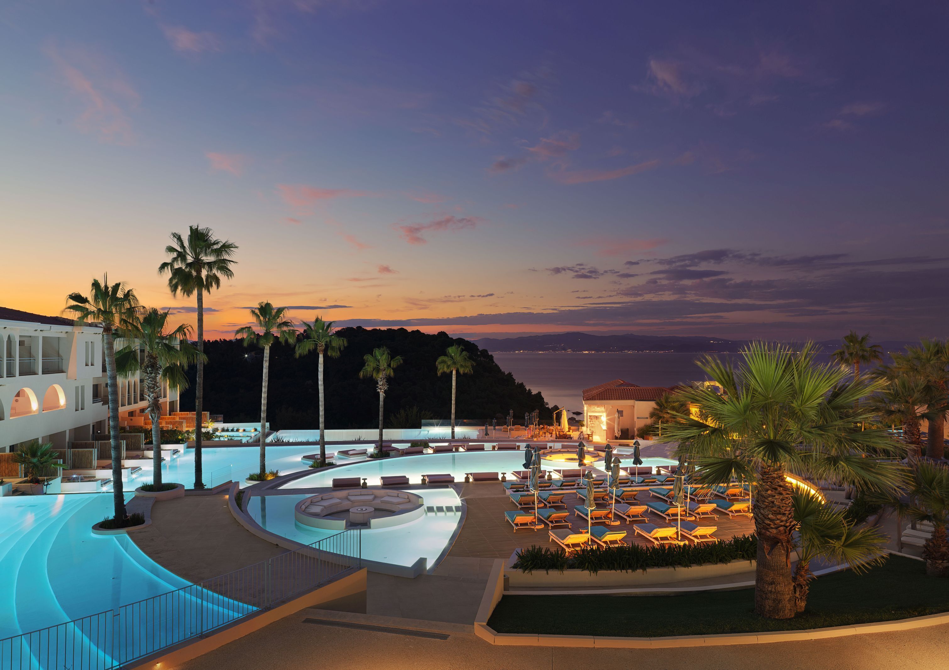 A resort with a large pool, palm trees, and a view of the sea at sunset.