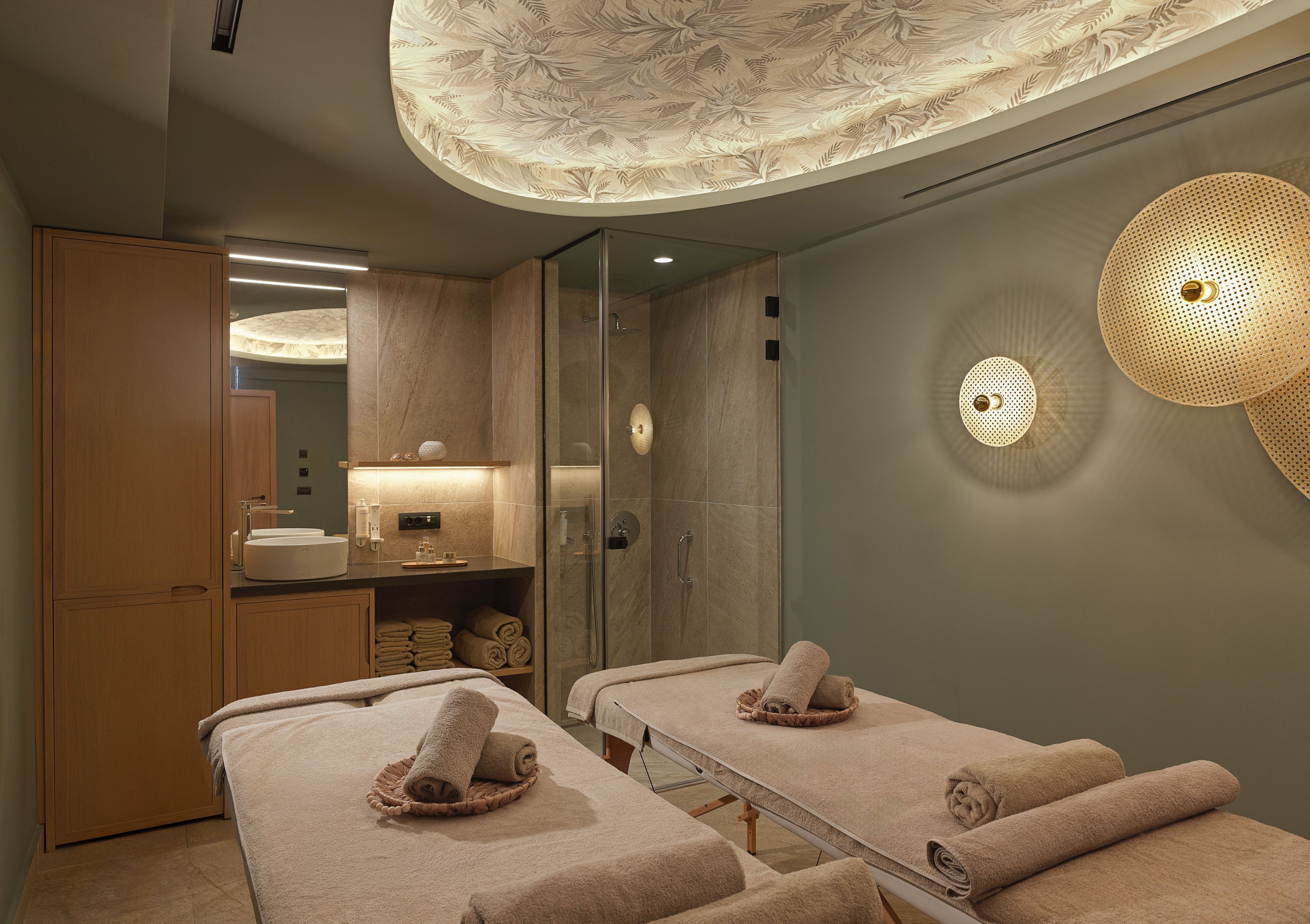 A spa room with two massage tables, a wooden cabinet, a mirror, and a ceiling with a leaf pattern.