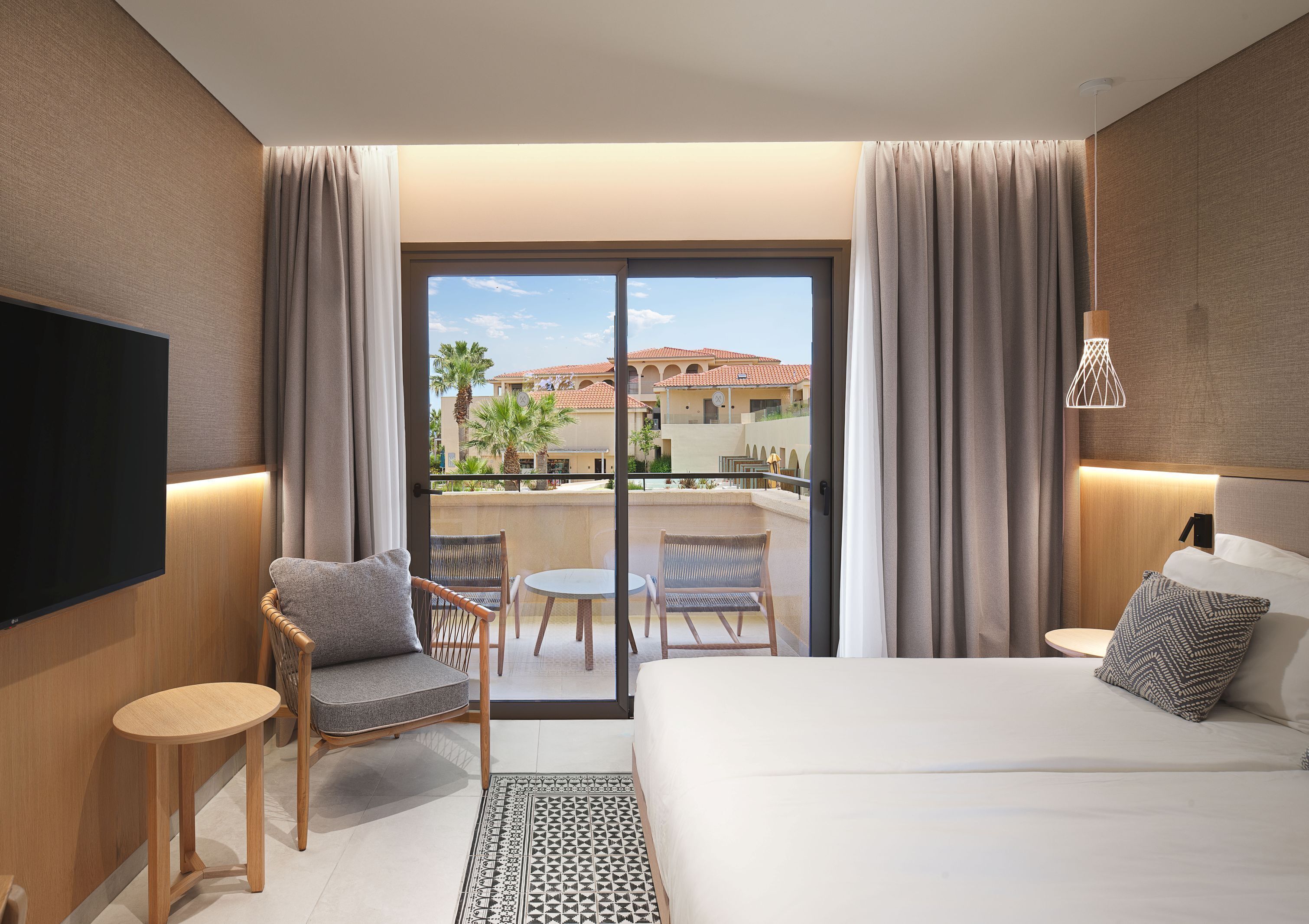 A hotel room with a bed, a TV, a balcony with a table and chairs, and a view of buildings and palm trees.