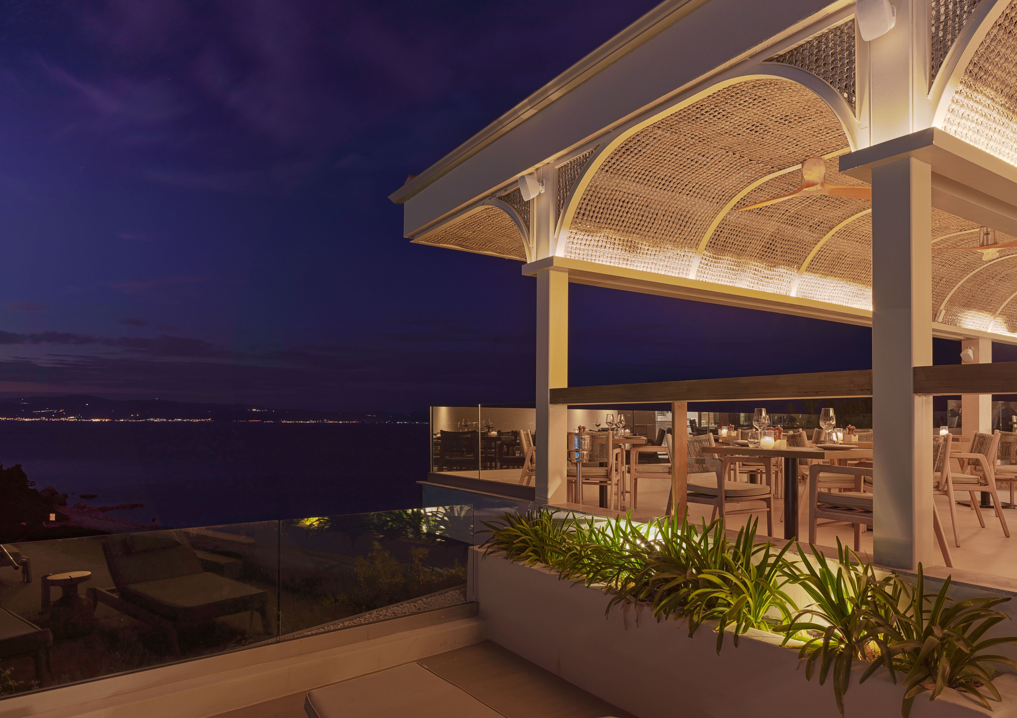 A modern outdoor dining area with a view of the sea, featuring a pergola with a woven ceiling and a seating arrangement.