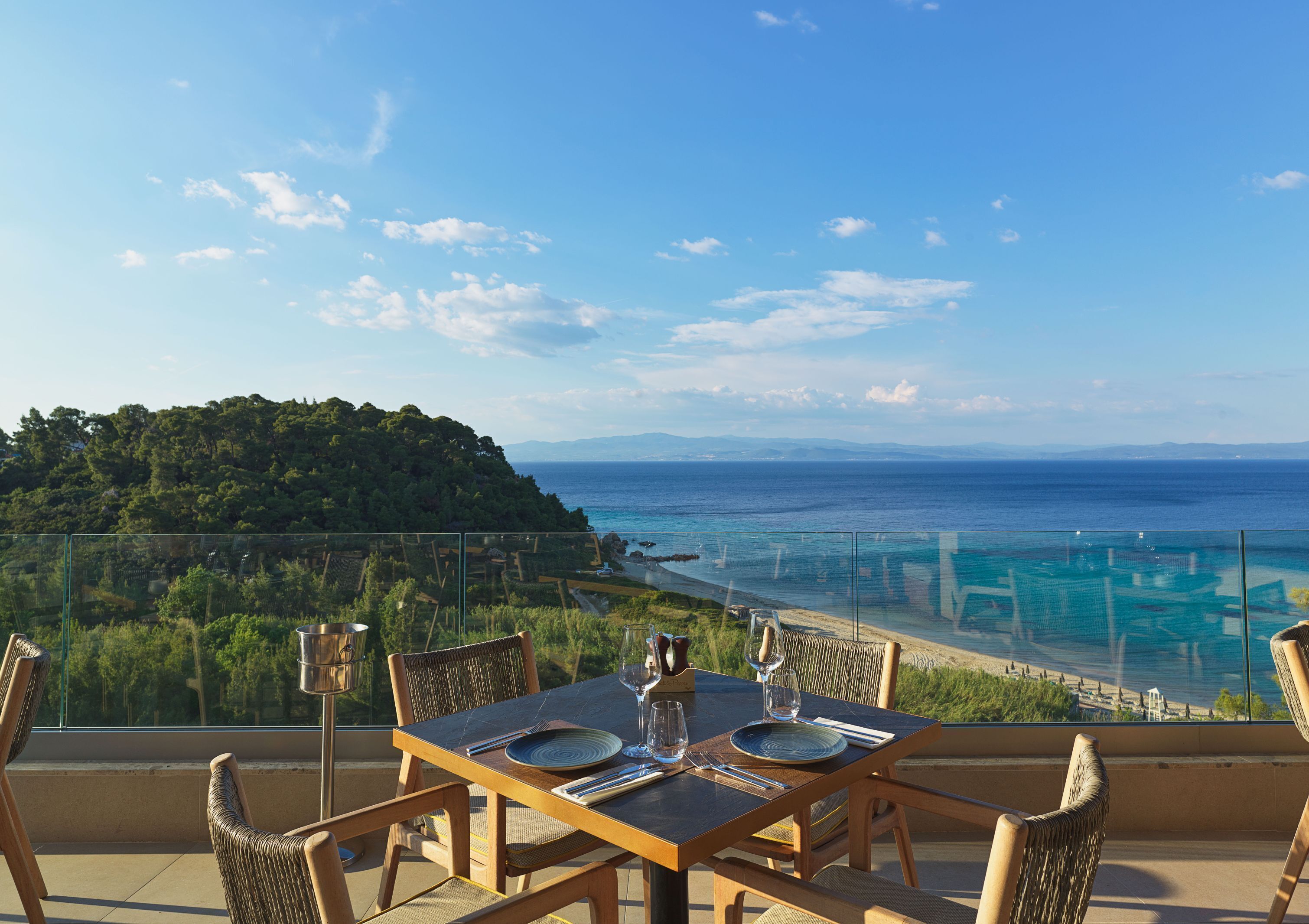 Une table à manger dressée avec des assiettes, des verres et des couverts, sur un balcon donnant sur une vue côtière, avec une colline boisée et un ciel bleu clair.