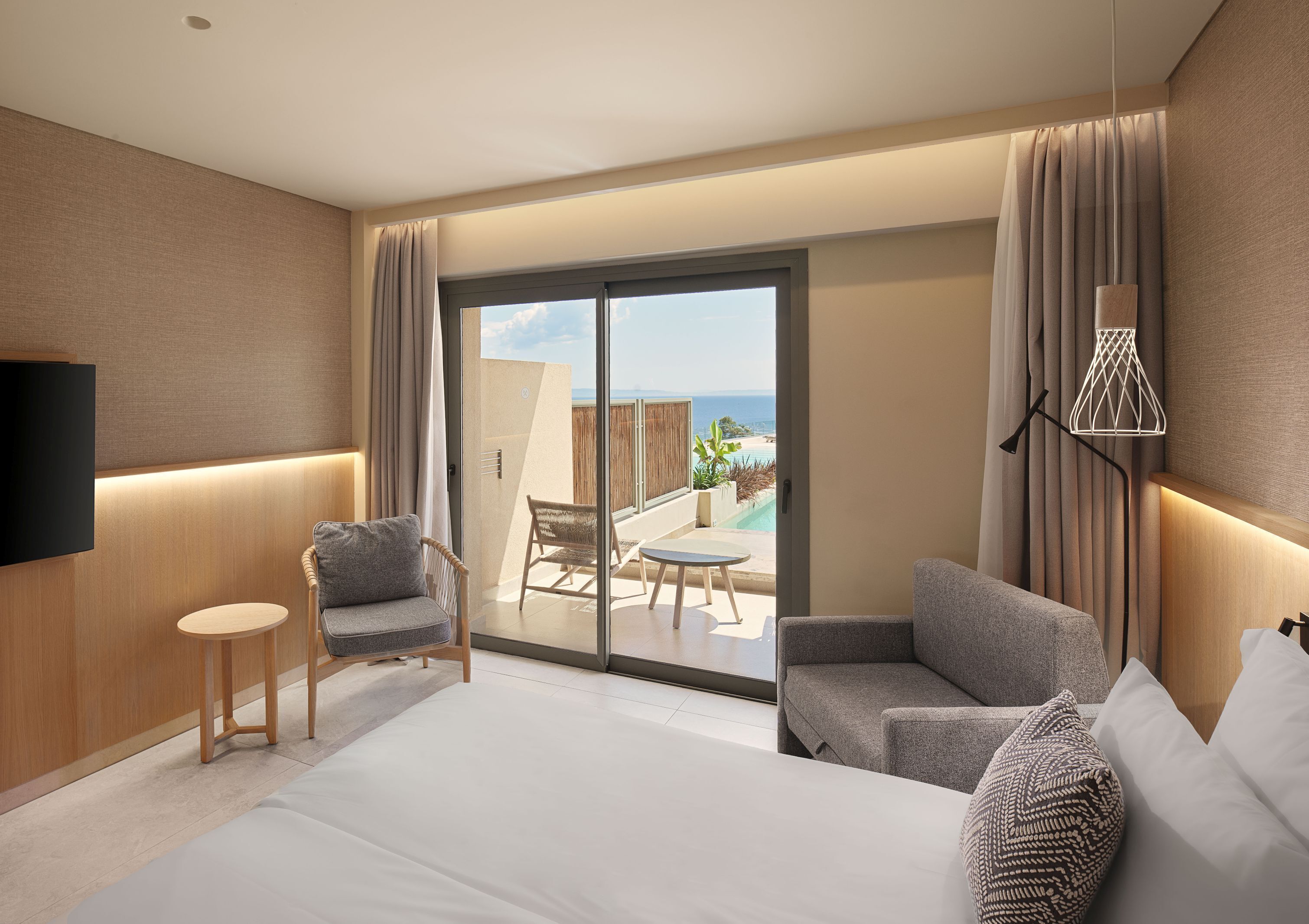 Habitación de hotel moderna con una cama grande, dos sillones, una mesita, un televisor y un ventanal corredero que da a un balcón con vistas a la piscina.
