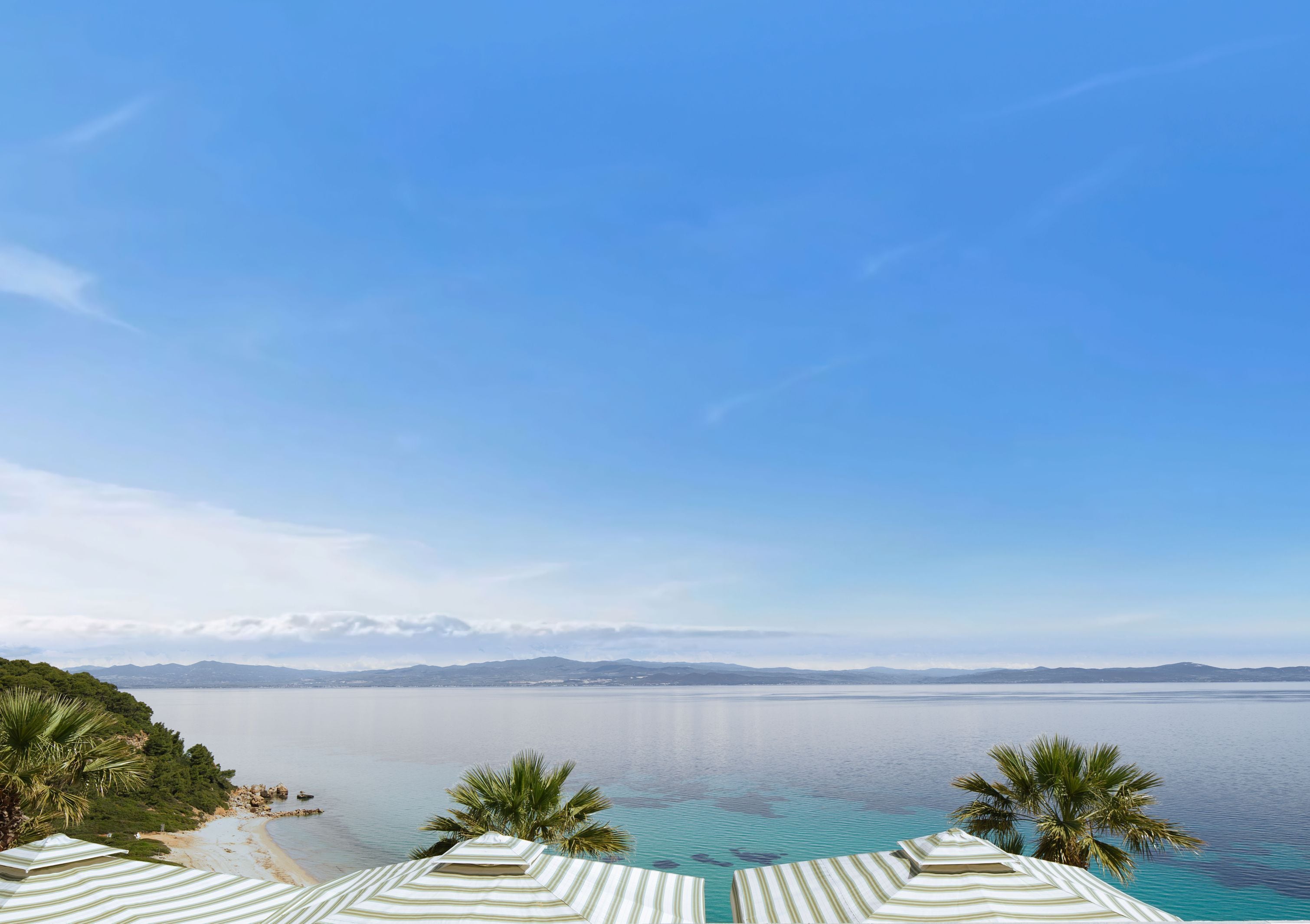 A beach view with a clear sky, a calm sea, and a mountain range in the distance.
