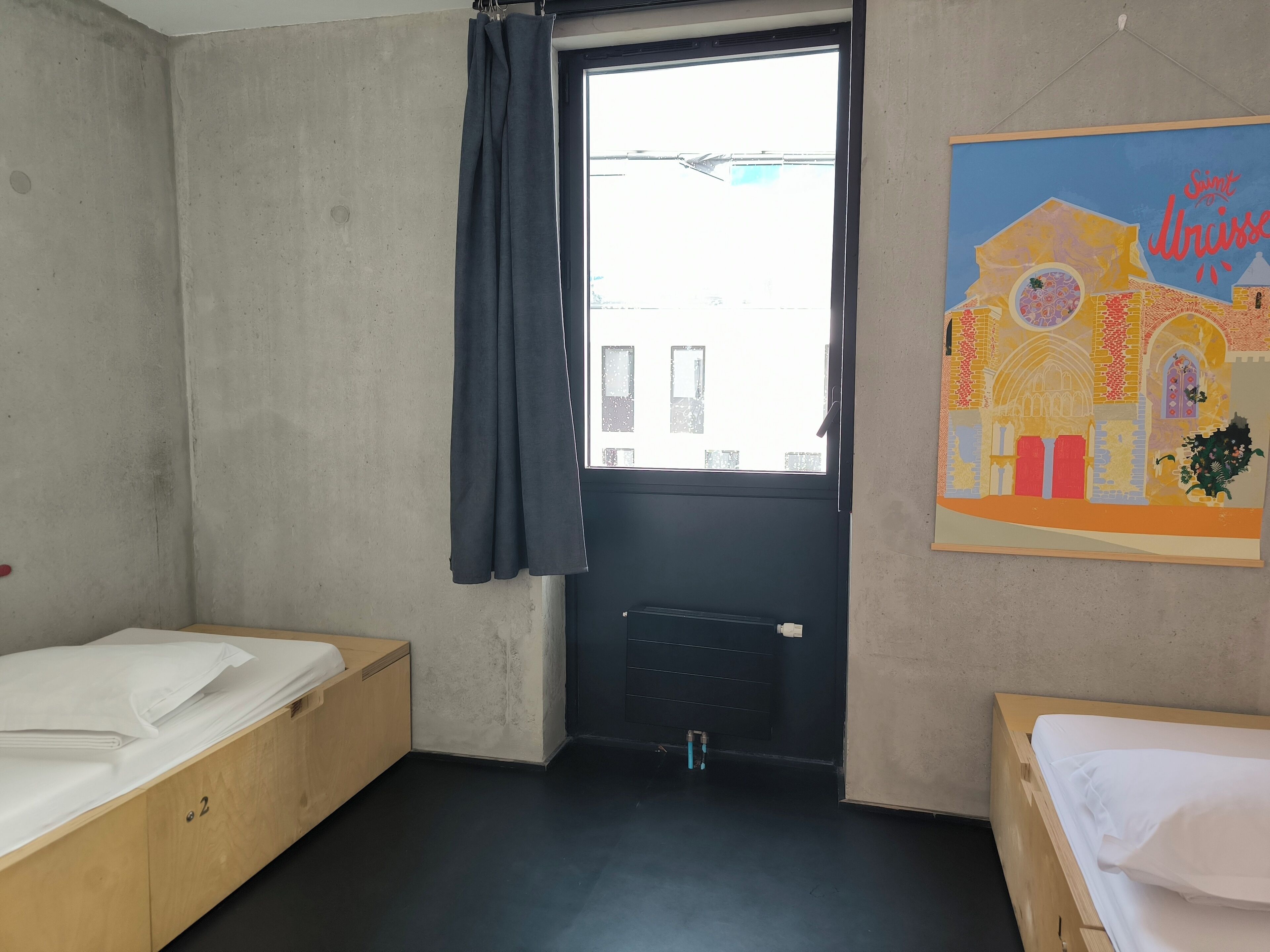 A room with two wooden beds, a window with a curtain, a radiator, and a colorful wall painting.