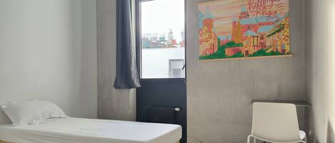 A bedroom with a bed, a chair, a window with curtains, and a cityscape painting on the wall.