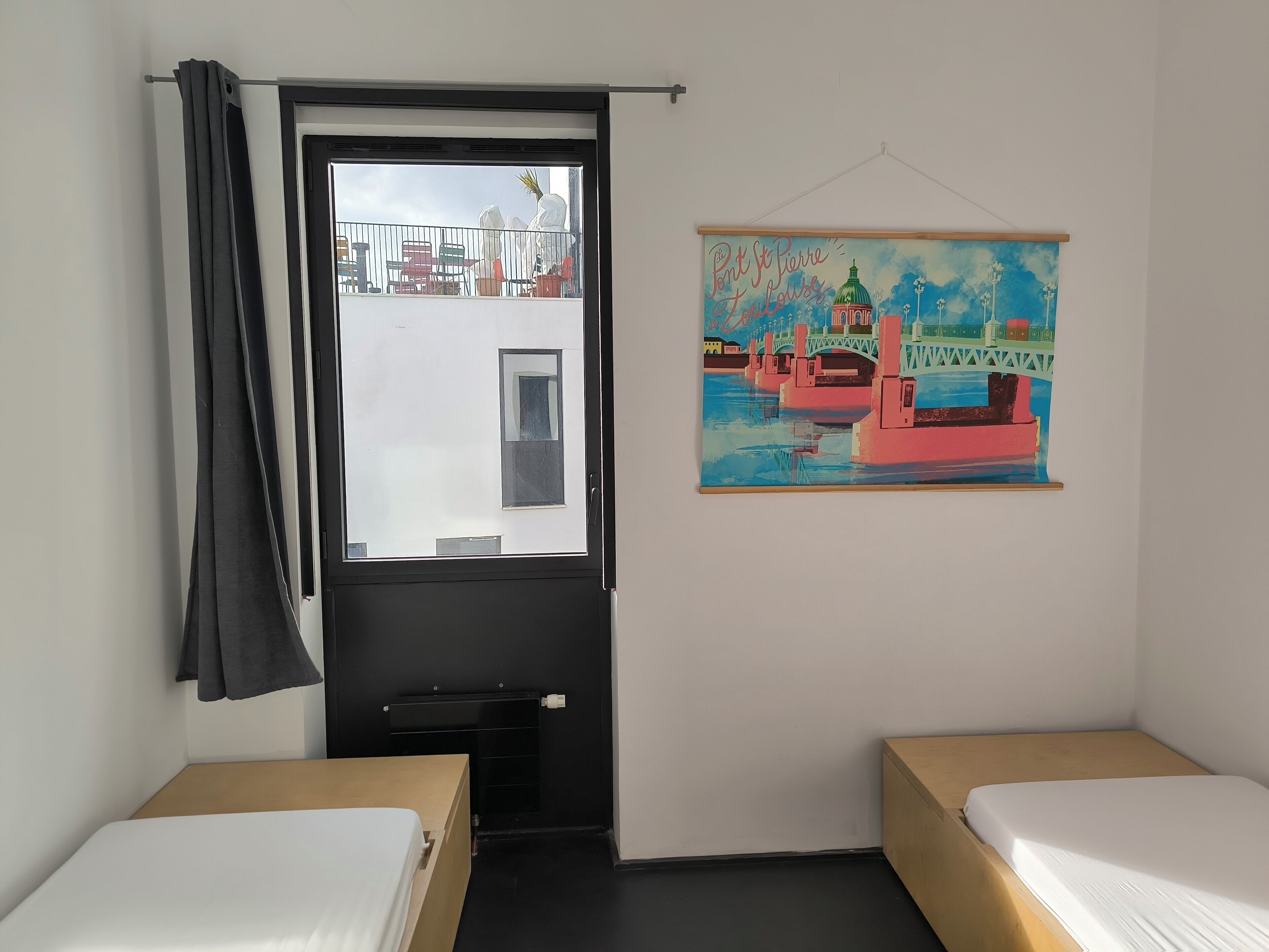 A room with two single beds, a black door, a window with a view of outdoor furniture, and a colorful painting of a bridge.