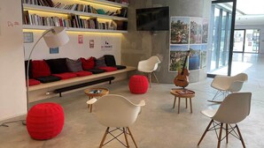 Un área de descanso moderna con sofá, pufs rojos, sillas blancas, una estantería y una guitarra.