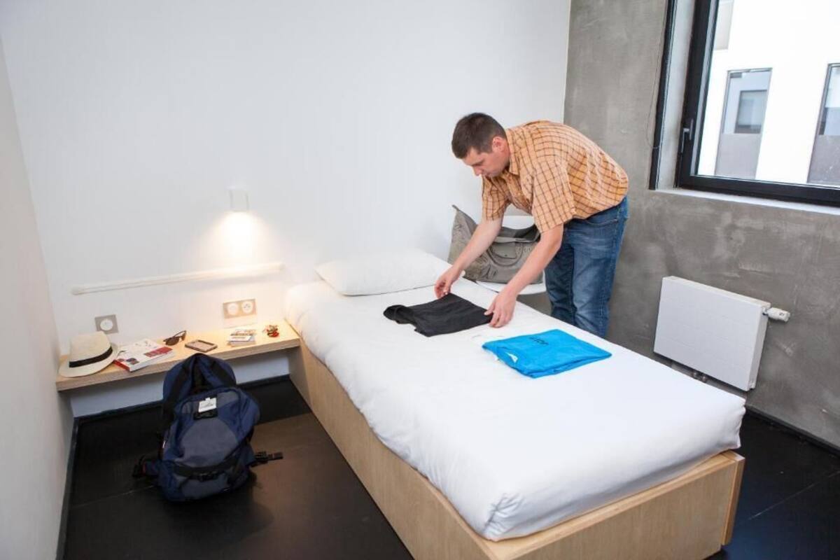 Un lit simple avec un matelas blanc et une serviette bleue, un sac à dos gris posé sur le sol, et une fenêtre donnant sur des immeubles.