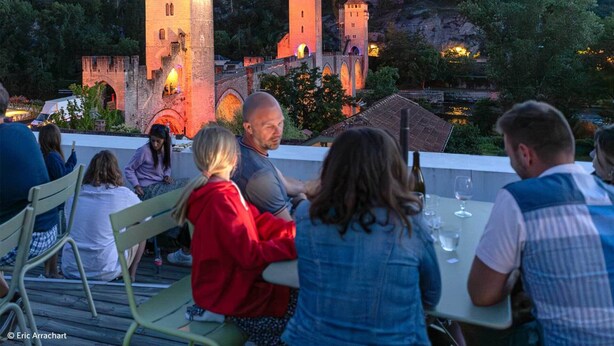 Un grupo de personas cenando en una mesa al aire libre con vistas a un castillo y vegetación circundante.