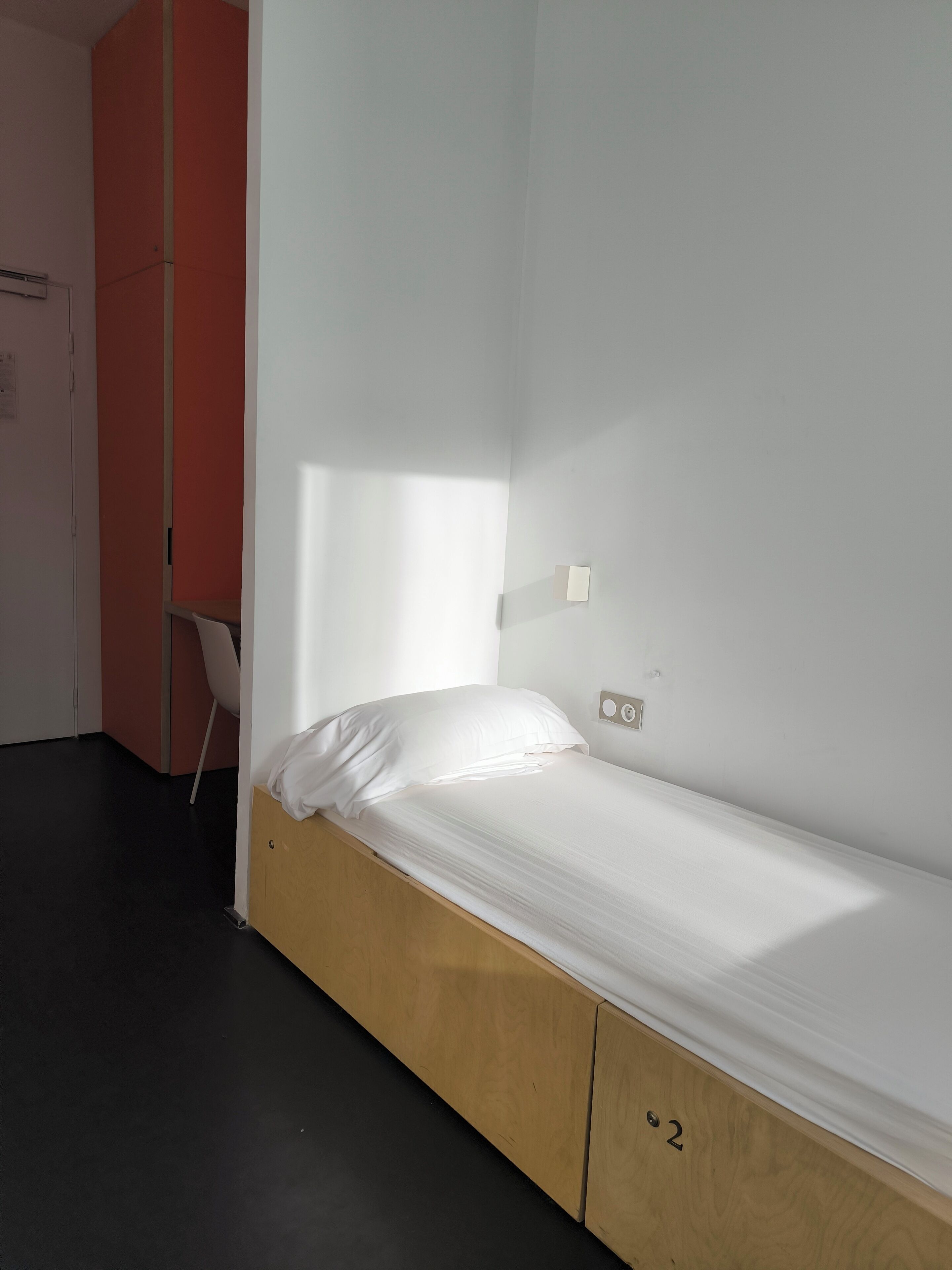 A single bed with a wooden frame and a white mattress in a room with a red partition and a white wall.