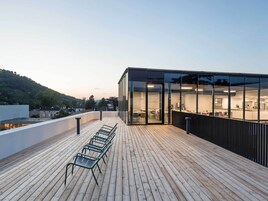 Un edificio moderno con fachada de vidrio, terraza de madera y zona de estar al aire libre.