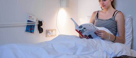 A person reading a book in a room with a bed, a wall-mounted light, and a suitcase.