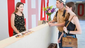 Un mostrador de recepción con un gran letrero rojo con la letra 'G', un florero con flores y una maleta en el suelo.