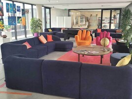 Un área de descanso moderna con sofás azul oscuro, un sillón naranja brillante y una mesa redonda con revistas.