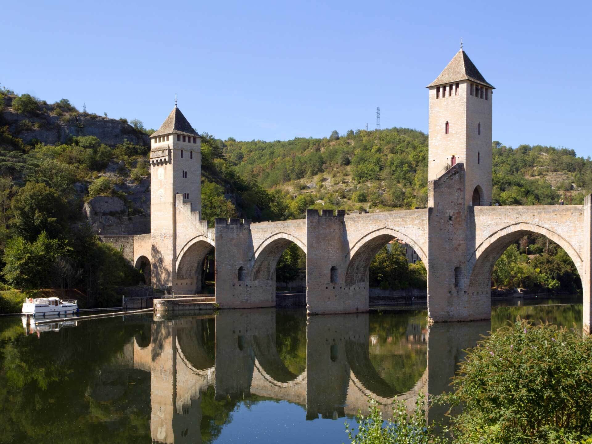 A stone bridge with towers and arches over a calm river, surrounded by greenery and hills.