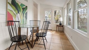 A dining area with a round glass table and black chairs, a colorful abstract painting on the wall, and a view through the window.