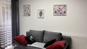 A modern living room with a grey sofa, red and black cushions, a patterned rug, and floral artwork on the walls. - Apartment with terrace, 48sqm. for max. 4 persons (Bühlertal)