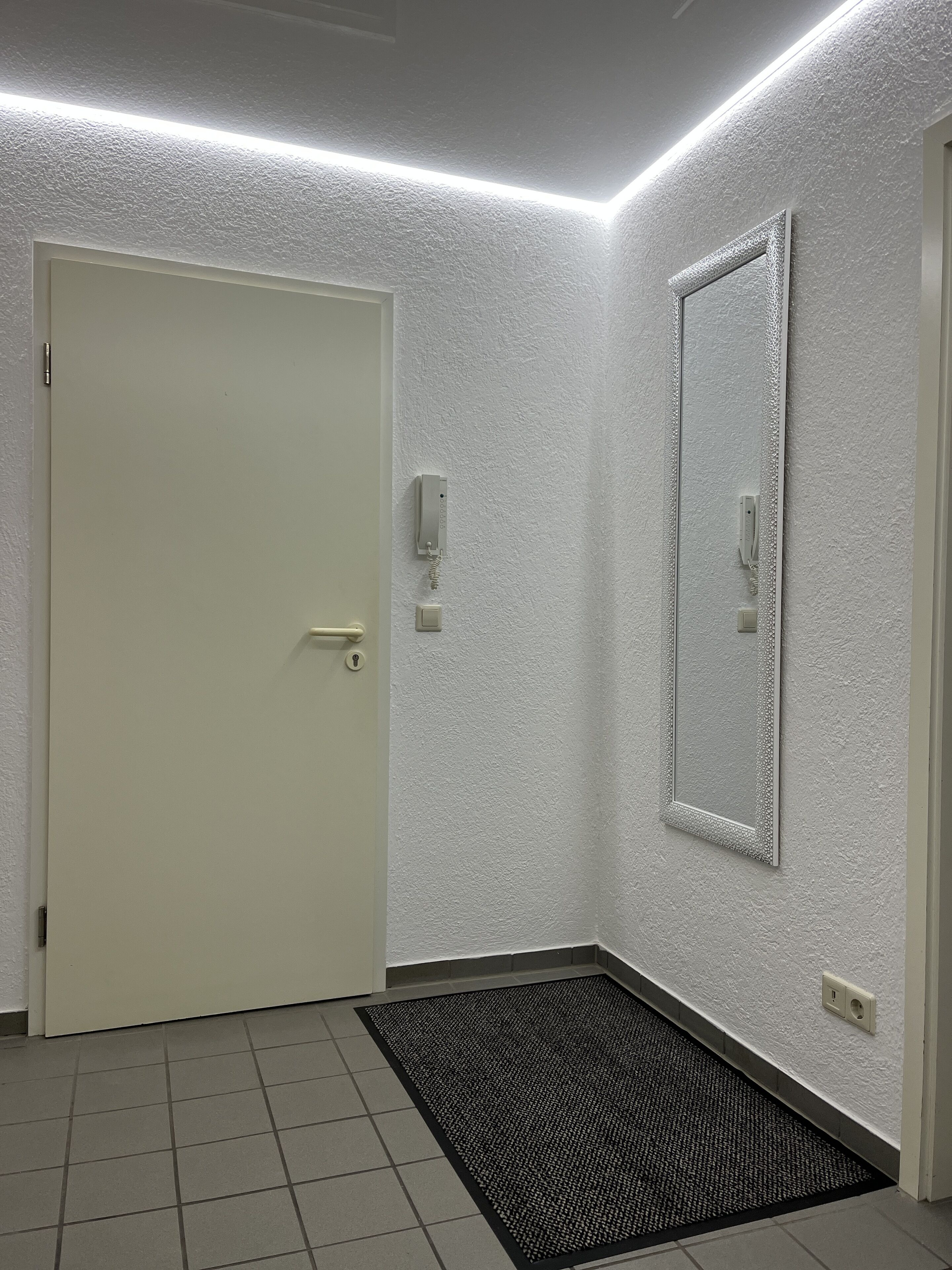 Een gang met een witte deur, een structuurwand, een spiegel en een zwarte deurmat.